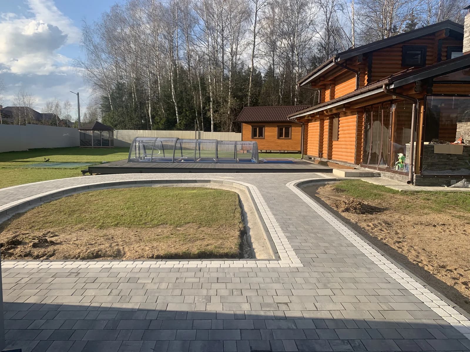 Задний двор с бассейном под навесом, газоном и серыми мощёными дорожками вокруг деревянного дома
