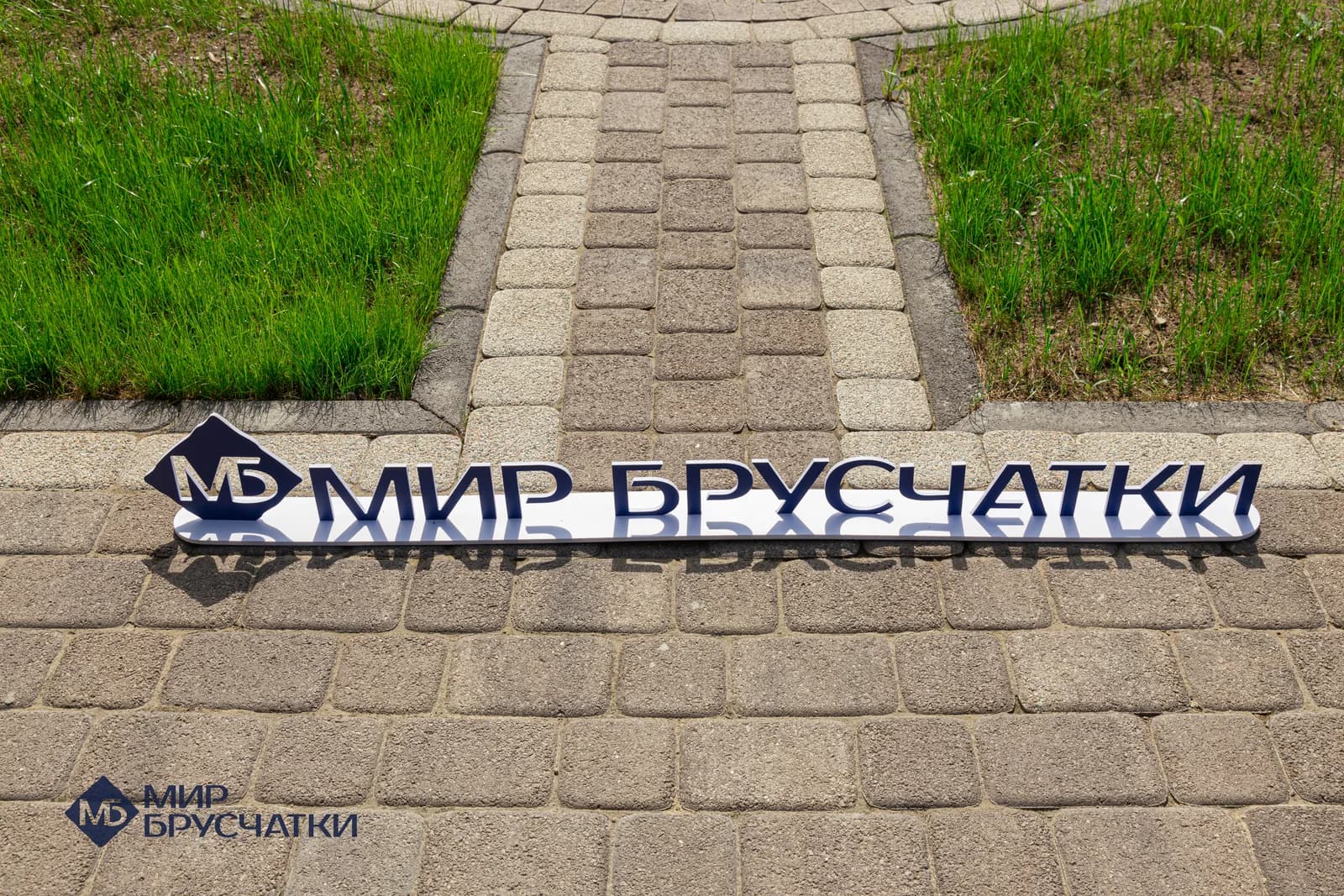Металлическая табличка «Мир Брусчатки» лежит на мощении крупным планом: брусчатка «старый город» серо-бежевого цвета, по краям трава.