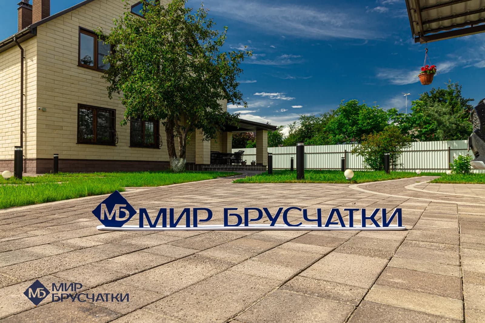 Табличка «Мир Брусчатки» стоит на вымощенном плиткой дворе перед жёлтым кирпичным домом с навесом.