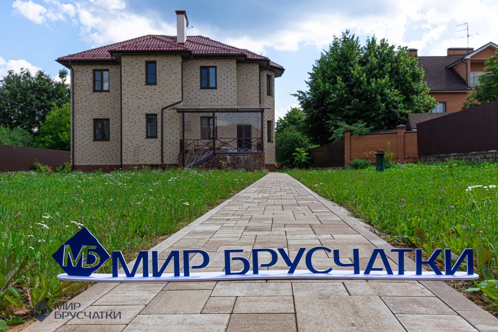 Дорожка из брусчатки к кирпичному дому через газон, на переднем плане логотип «Мир Брусчатки»