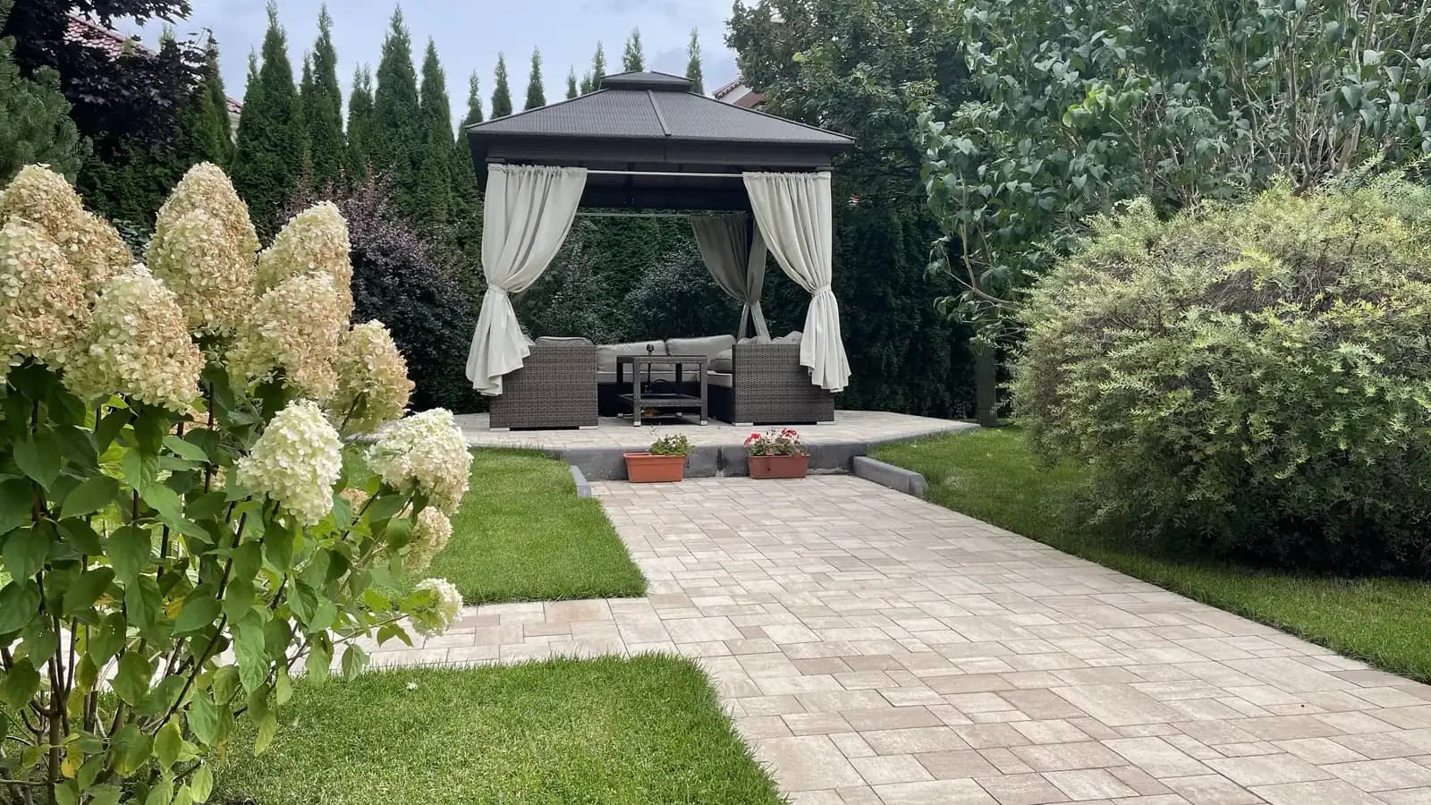 Беседка с мягкой мебелью и мощёная дорожка из бежевой плитки в озеленённом саду с гортензиями